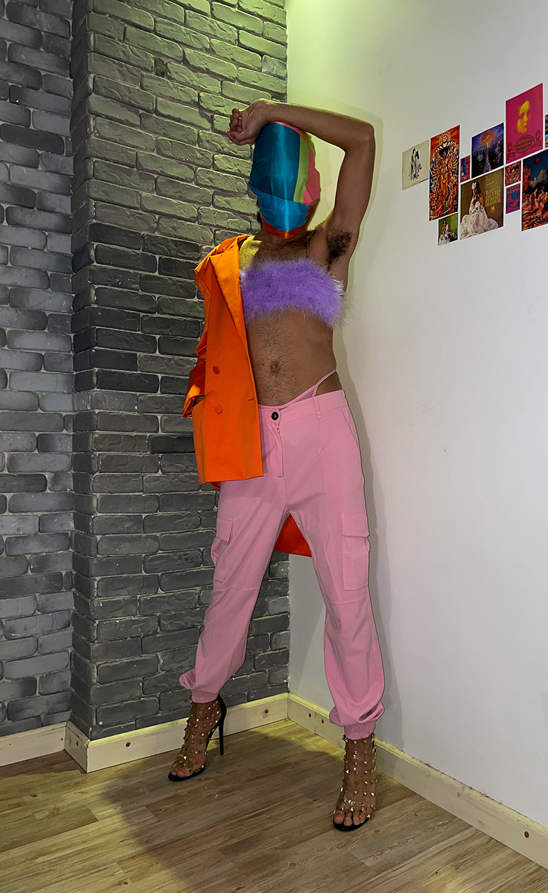 A person stands defiantly with their face covered in a mutlicolor silk fabric. They're wearing a furry purple bralet, pink cargo pants with a pink thong poking out, bedazzled high heels, and an orange coat that is slung on one shoulder. They're standing in the corner of two walls—one is a gray brick and the other is painted white.
