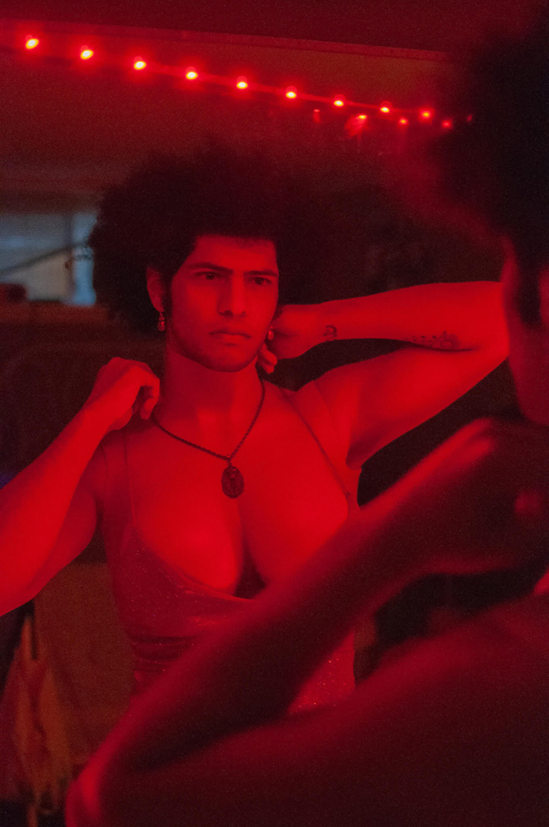 A person with an afro and facial hair wearing a glittery, low-cut dress looks intensly into a mirror. They are bathed in red light and are touching a necklace around their neck with a large oval pendant. 