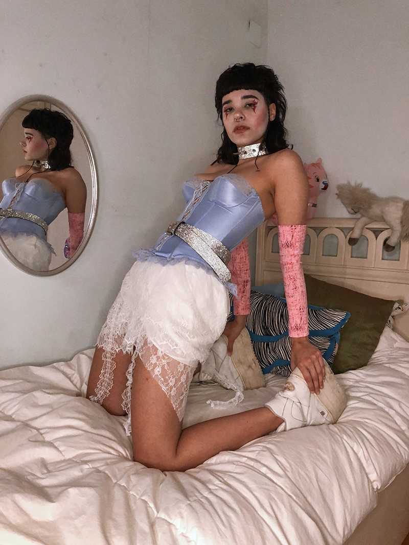 A femme person with black hair that is styled into a mullet with short bangs. They have white make up on their face with red lines running downward over their eyes. They are perched on a bed on their knees with their back stretched slightly backward and their hands grabbing the back of their ankles. They wear a light blue silky corset, a short white lace skirt, and white boots with large platforms. They are adorned with a matching silver glitter belt and choker.